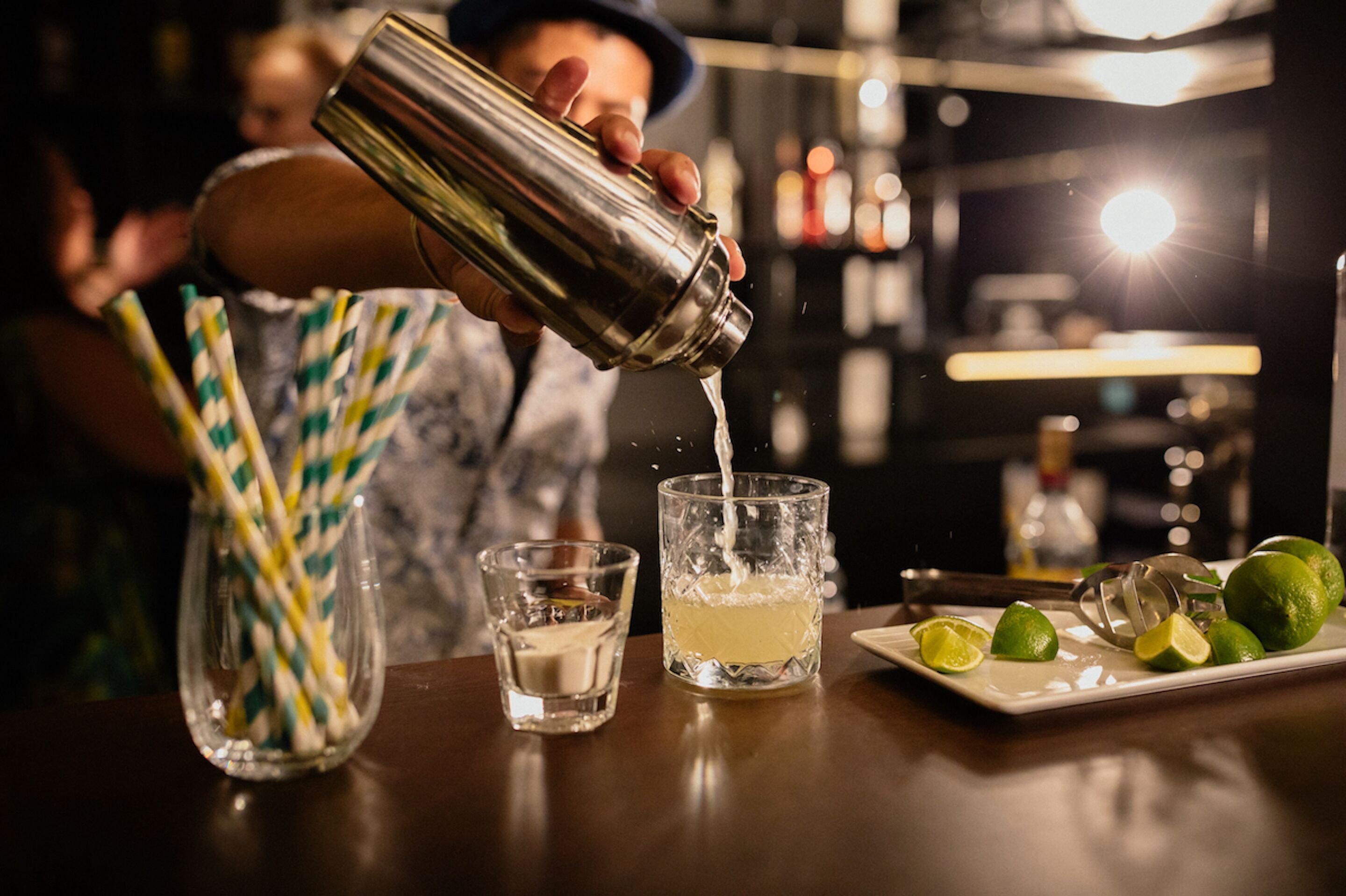 A bartender pouring a drink into a glass, with a tray of limes and straws nearby.