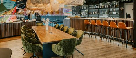 Zona de bar moderna con mostrador de madera, diversas opciones de asiento como taburetes y sillas acolchadas, y una variedad de botellas y vasos en estantes.