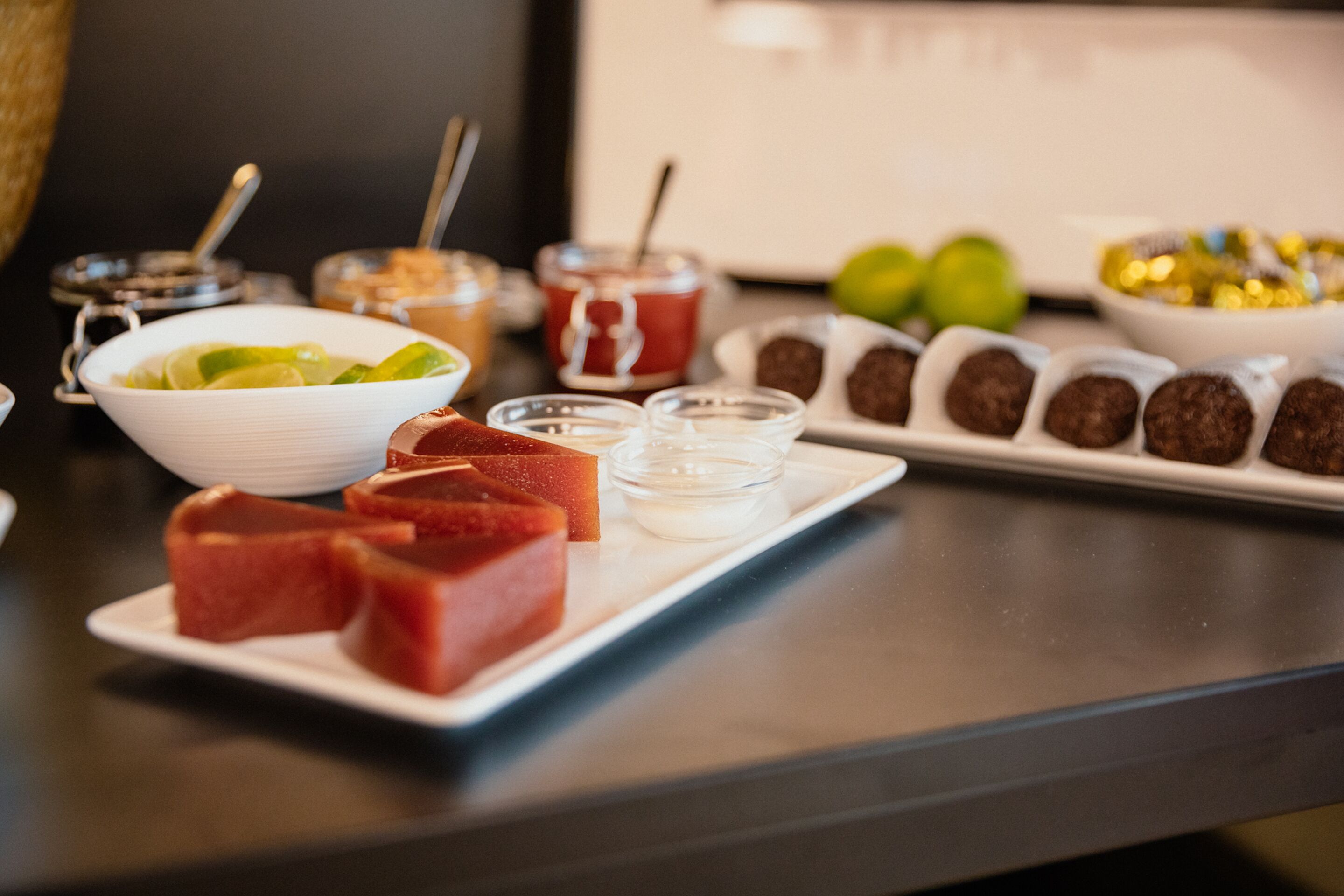 Una mesa con diversos alimentos, incluyendo un bol blanco con rodajas de lima, un plato con cubos rojos y una bandeja con bolitas de chocolate.