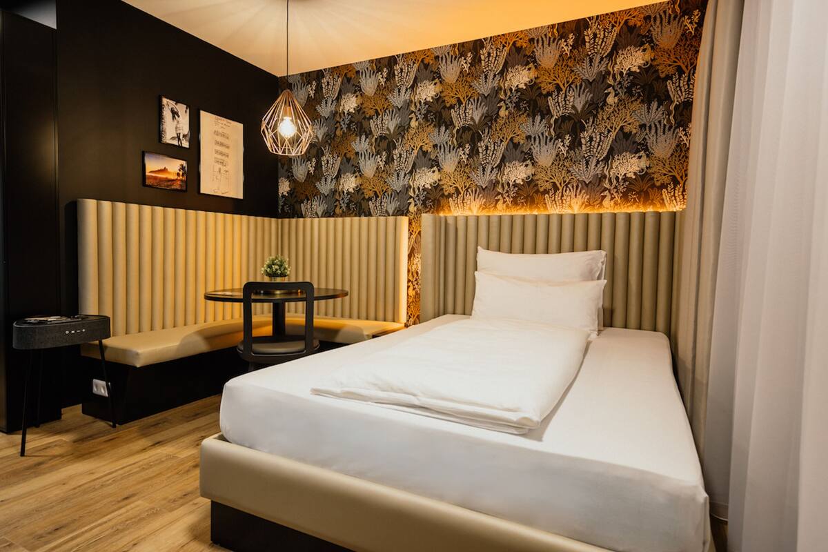 A modern hotel room with a bed, a bench, a small table, and a wall with patterned wallpaper.