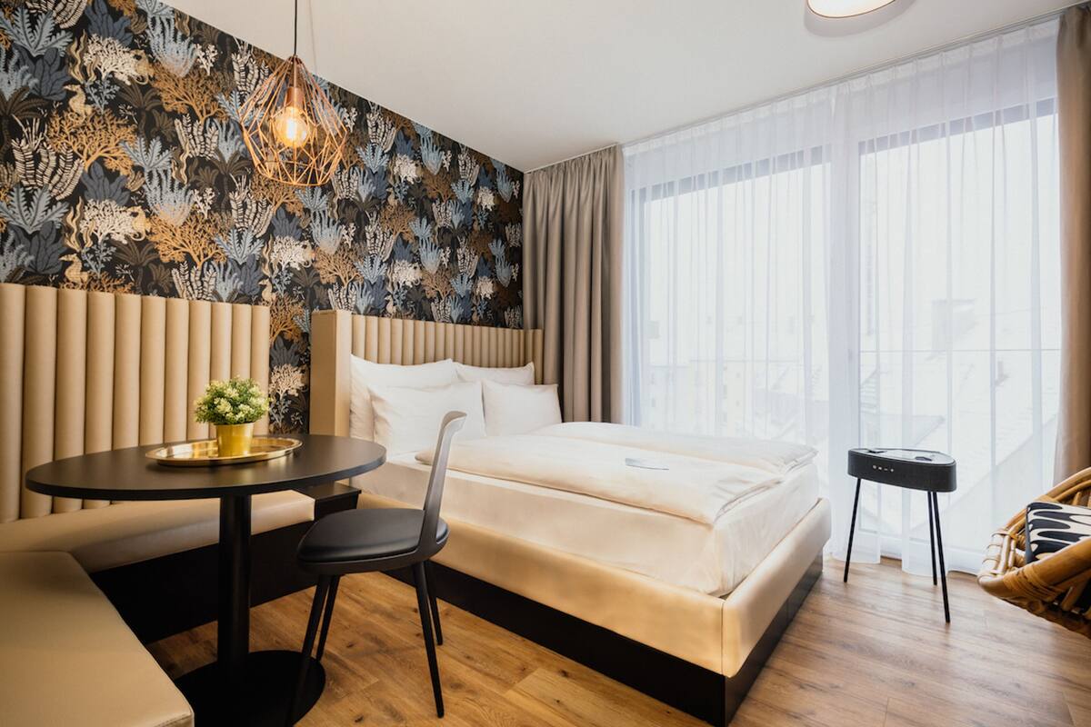 A modern hotel room with a large bed, a round table, a chair, a small side table, and a decorative wall with a geometric light fixture.