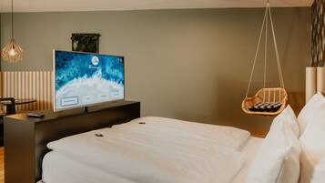 A modern hotel room with a bed, a television displaying a scenic wallpaper, a hanging chair, and a wooden side table.