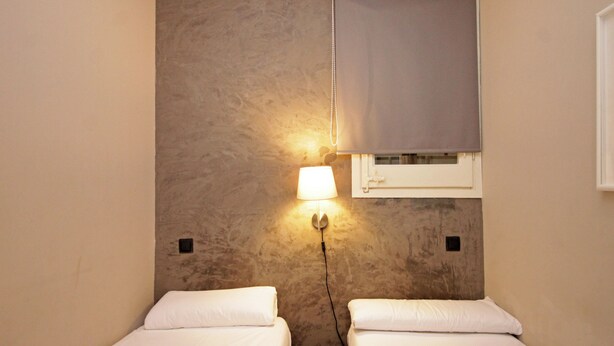A small, cozy room with two single beds, a wall-mounted lamp, and a window with a blind.