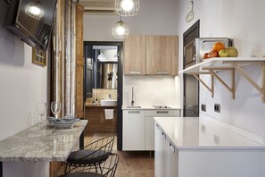 A modern kitchen with white cabinetry, a marble countertop, and a built-in oven. There is a wooden shelf with fruits and a clock on the wall.