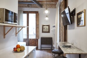 A compact kitchen with a dining area, wooden beams on the ceiling, a TV mounted on the wall, and a fruit bowl on the counter. - Studio with balcony, equipped with kitchenette and bathroom (Barcelona)