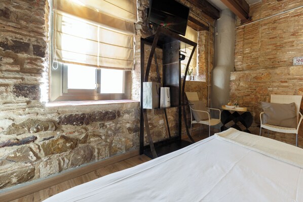 A rustic room with stone walls, a bed, a TV mounted on a metal frame, and a window with blinds.