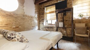 Une pièce au style rustique, avec des murs en briques apparentes, un lit agrémenté d’oreillers à motifs, une chaise en osier et une fenêtre avec des rideaux.