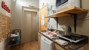 Une cuisine compacte équipée d’un four à micro-ondes, d’une bouilloire et d’un petit évier. Au-dessus du plan de travail, on trouve une étagère en bois et, en dessous, un porte-vaisselle en métal.