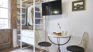Une pièce avec une armoire blanche, un téléviseur fixé au mur, une petite table ronde avec deux chaises et un tableau encadré accroché au mur.