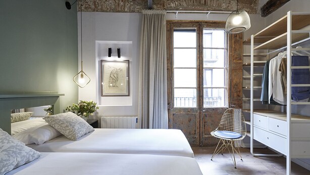 Une chambre avec deux lits, une grande fenêtre avec des rideaux, un radiateur, un tableau encadré, un luminaire suspendu et une armoire.