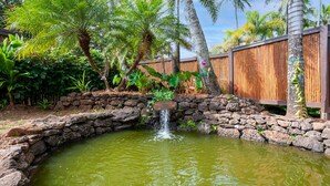 Un petit étang avec une cascade, entouré de plantes tropicales et d’une clôture en bois.