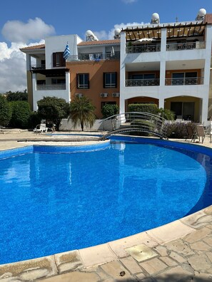 A swimming pool with a stone border, a covered patio with outdoor furniture, and a multi-story building with balconies and satellite dishes.