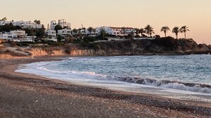 A coastal view with a beach, rocky shoreline, and white buildings with palm trees. - 2 Bedroom Ground Floor Apartment Close to Shops, Restaurants, Old Town and beach (Paphos)