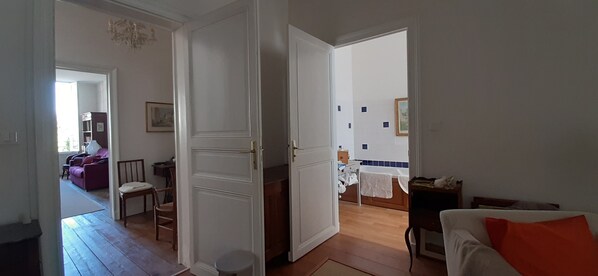 A room with a wooden floor, a white door, a bed with a white cover, a small table, and a chair.