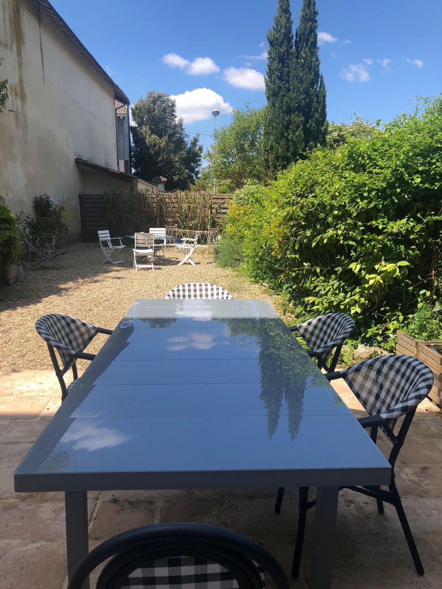 Patio esterno con tavolo rettangolare e quattro sedie, circondato da piante e un albero alto.
