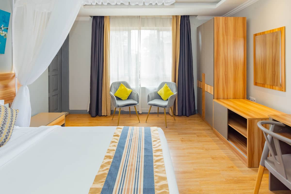 A hotel room with a bed, a desk, two chairs, a wardrobe, and a large window with curtains.