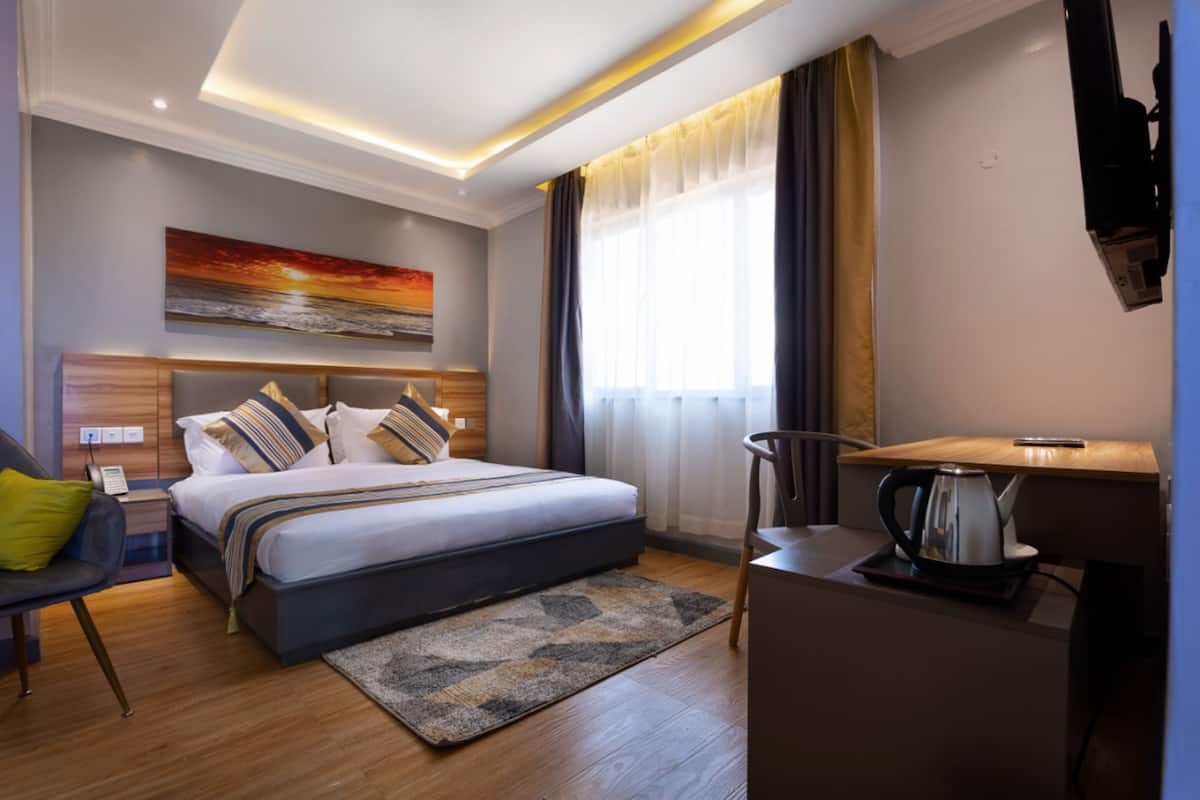 A modern hotel room with a large bed, a desk with a kettle, a chair, and a painting on the wall.