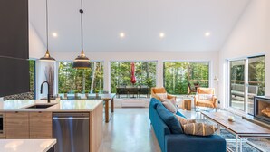 Eine moderne Küche mit Geschirrspüler, Essbereich mit Tisch und Stühlen, ein blaues Sofa und große Fenster mit Blick auf Bäume.