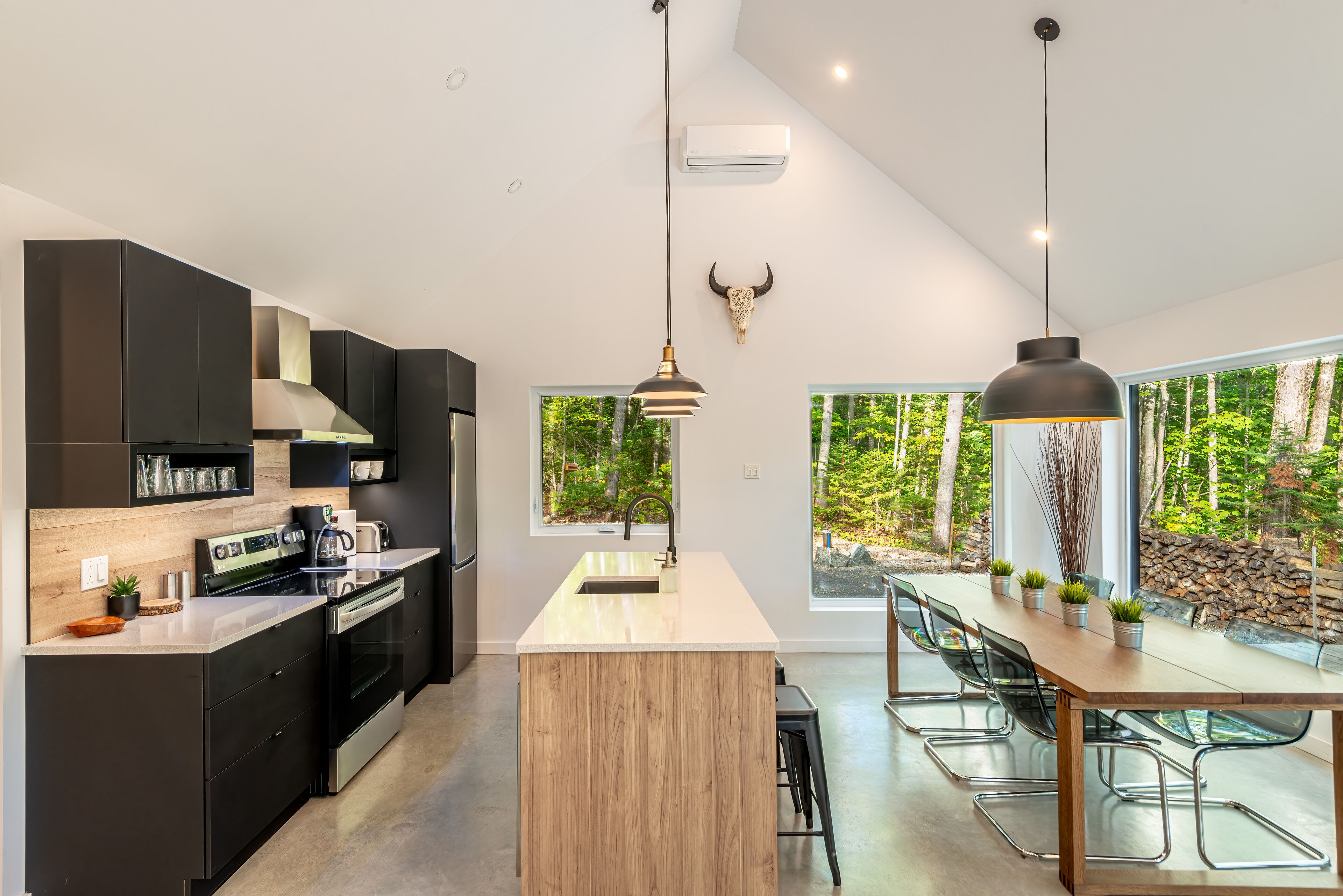 A modern kitchen with a central island, black cabinets, stainless steel appliances, and a large window overlooking a forest.
