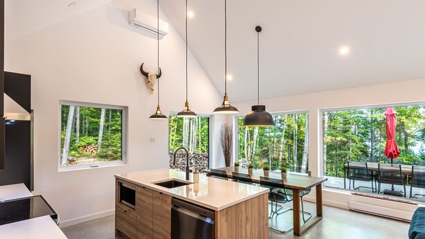Eine moderne Küche mit einer großen Kochinsel, Pendelleuchten und Blick auf den Wald durch bodentiefe Fenster.