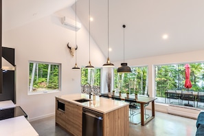 Een moderne keuken met een groot eiland, hanglampen en uitzicht op het bos door grote ramen.