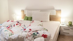 Un lit bien fait, recouvert d’une couette à motifs floraux, avec des oreillers blancs et une serviette verte. On y trouve une table de chevet avec une lampe et un petit objet décoratif.