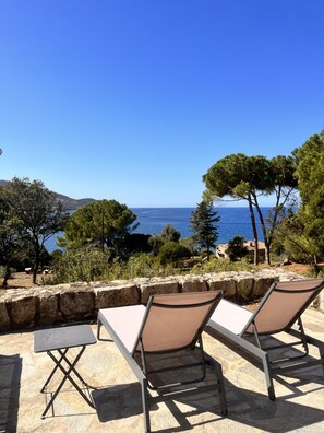 A terrace with two lounge chairs and a table, overlooking a coastal landscape with trees and a clear blue sky.