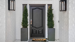 Ingresso moderno con una porta scura, due alberi sempreverdi in vaso e un tappetino con la scritta “HELLO”.