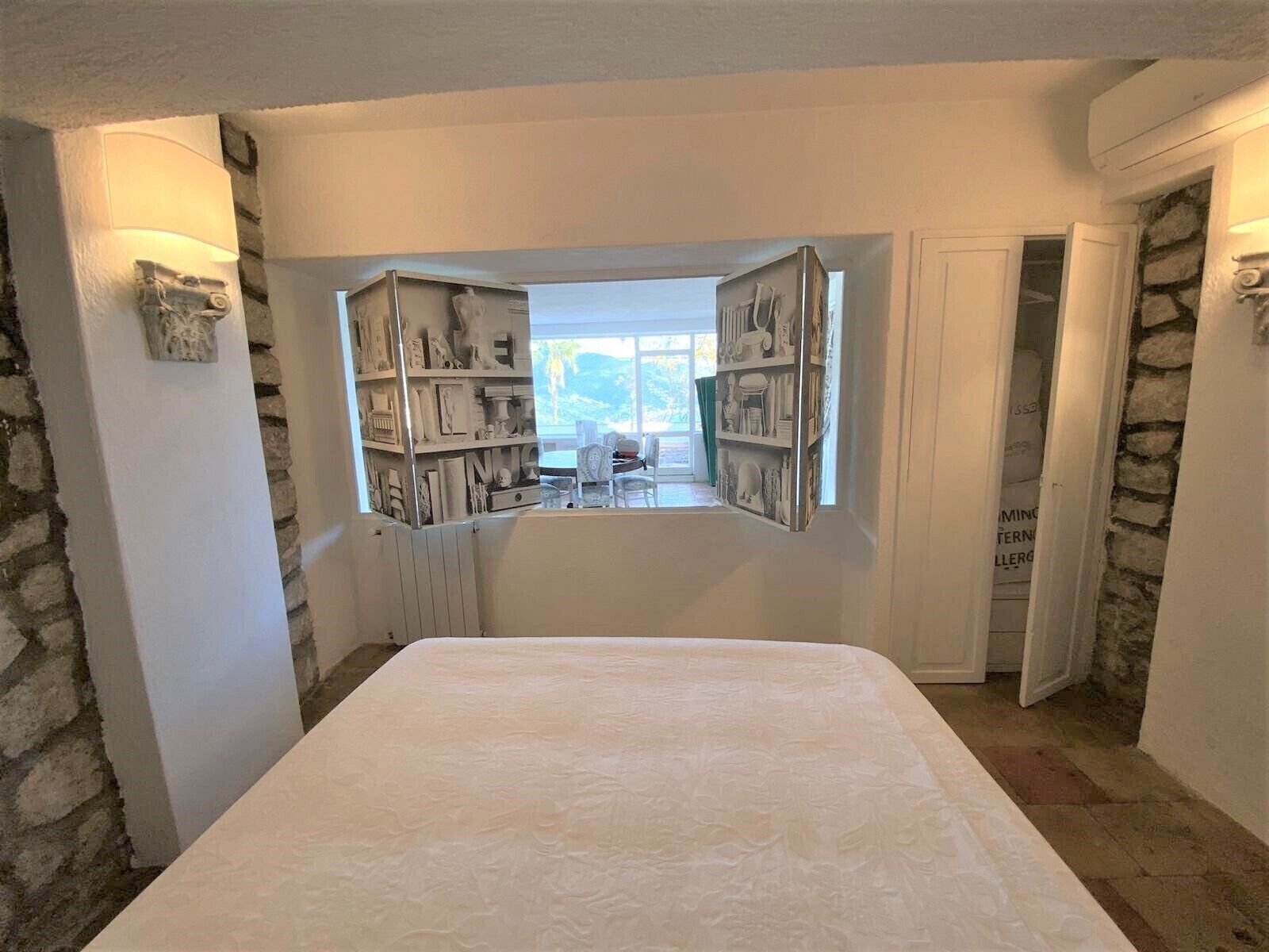 A bedroom with a large bed, a built-in bookshelf, a stone wall, and a view through a window.