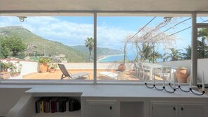 Een kamer met een groot raam met uitzicht op een kustlandschap, een boekenkast en een tafel met stoelen.