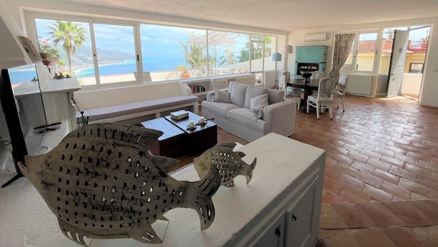 Een ruime woonkamer met een groot raam met uitzicht op zee, een witte bank, een eettafel en een decoratief beeld van een vis.