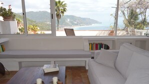 Een kamer met een witte bank, een houten tafel met boeken en kaarsen, een bankje en uitzicht op het strand en de bergen door grote ramen.