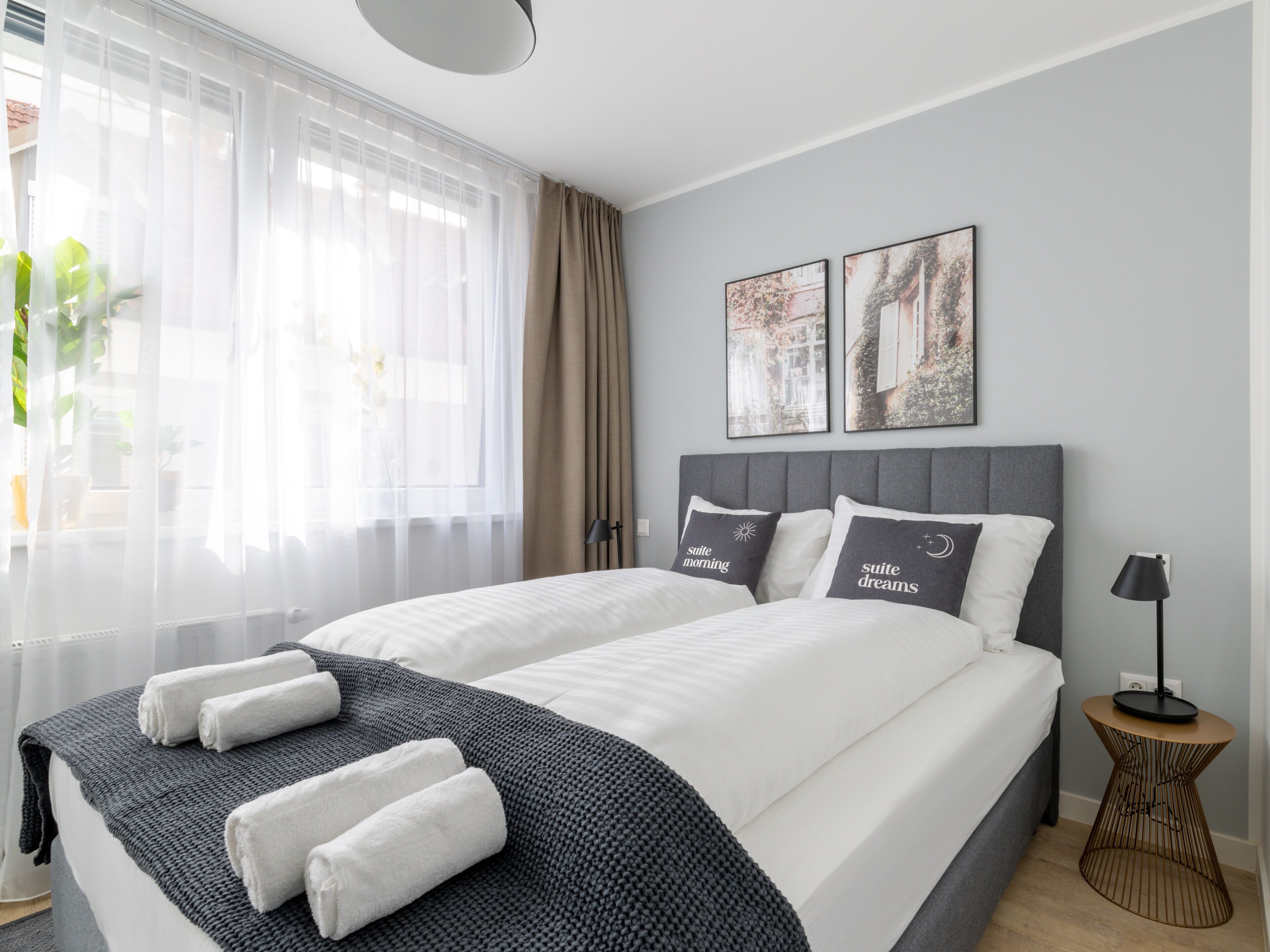 Habitació d'hotel moderna amb un llit gran, dos almoinos amb text, una taula auxiliar i una finestra amb cortines de tul.