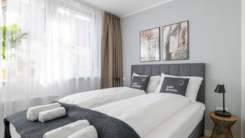 Habitació d'hotel moderna amb un llit gran, dos almoinos amb text, una taula auxiliar i una finestra amb cortines de tul.