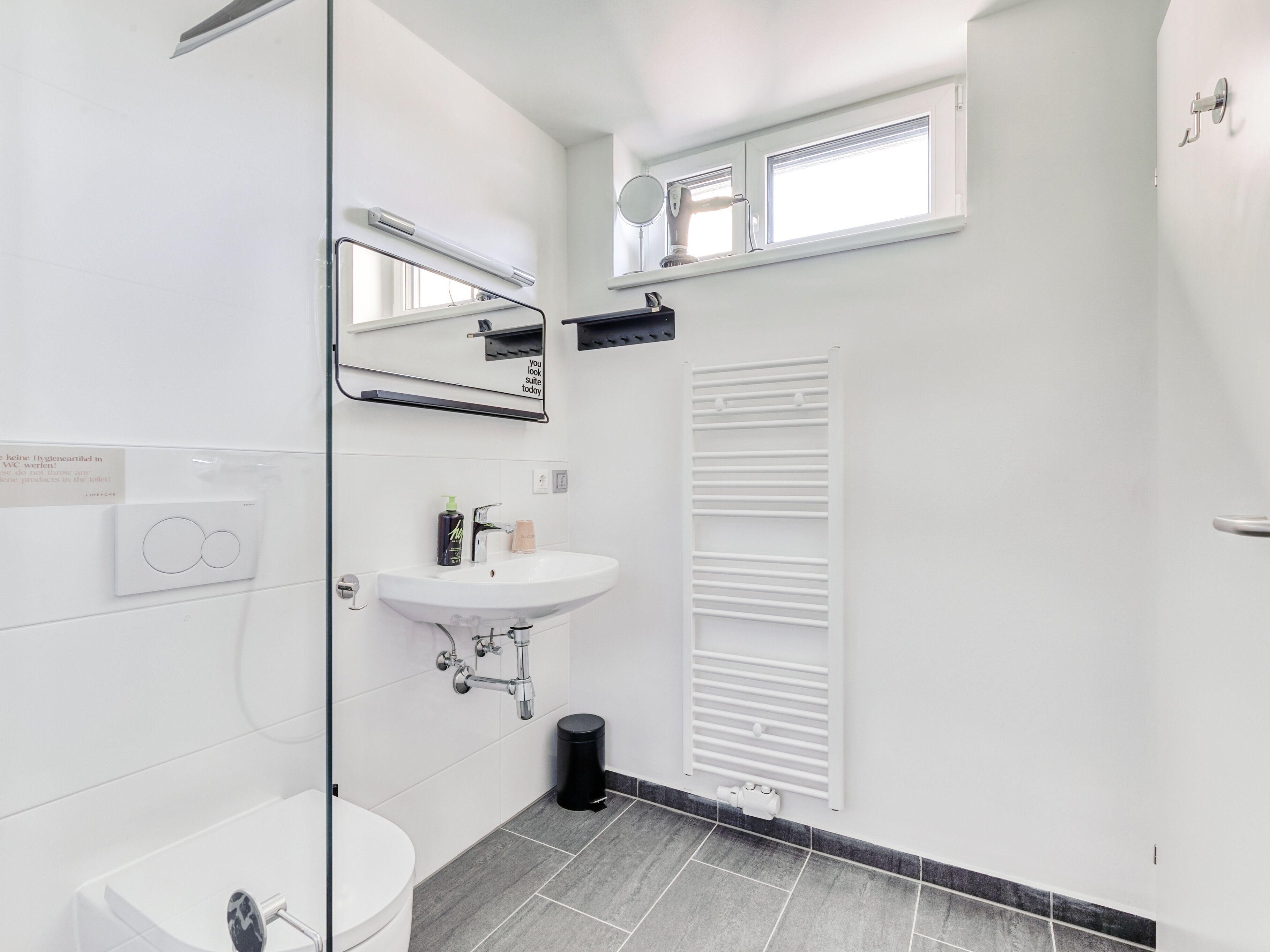Casa de banho moderna com lavatório branco, espelho e sanita. Tem uma janela com estores, um porta-toalhas e um lixeira preta.