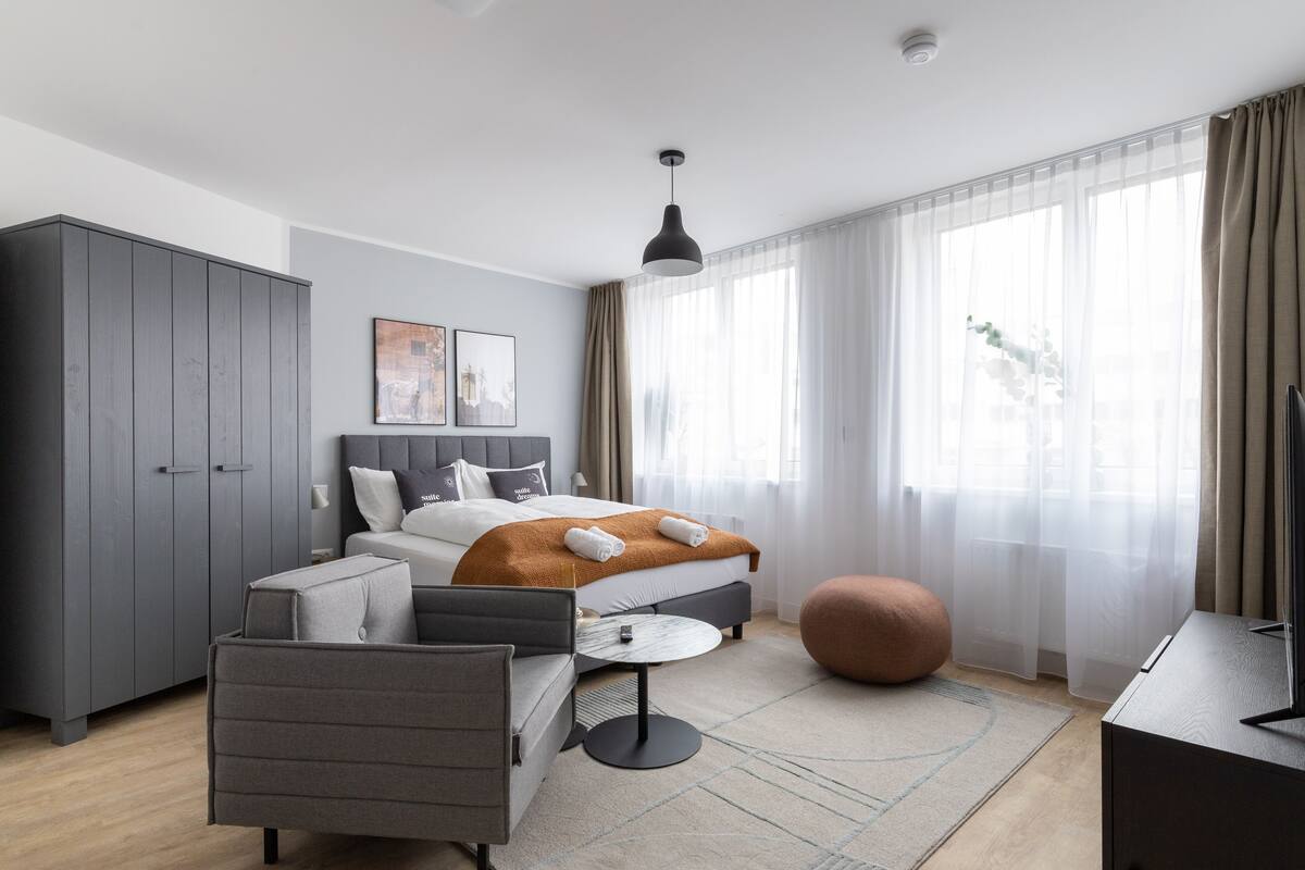 Une chambre d’hôtel moderne comprenant un lit, une armoire grise, un fauteuil, une table ronde, un pouf et un téléviseur.