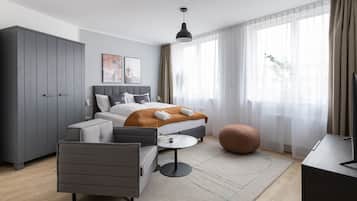 A modern hotel room with a bed, grey wardrobe, armchair, round table, ottoman, and a TV.