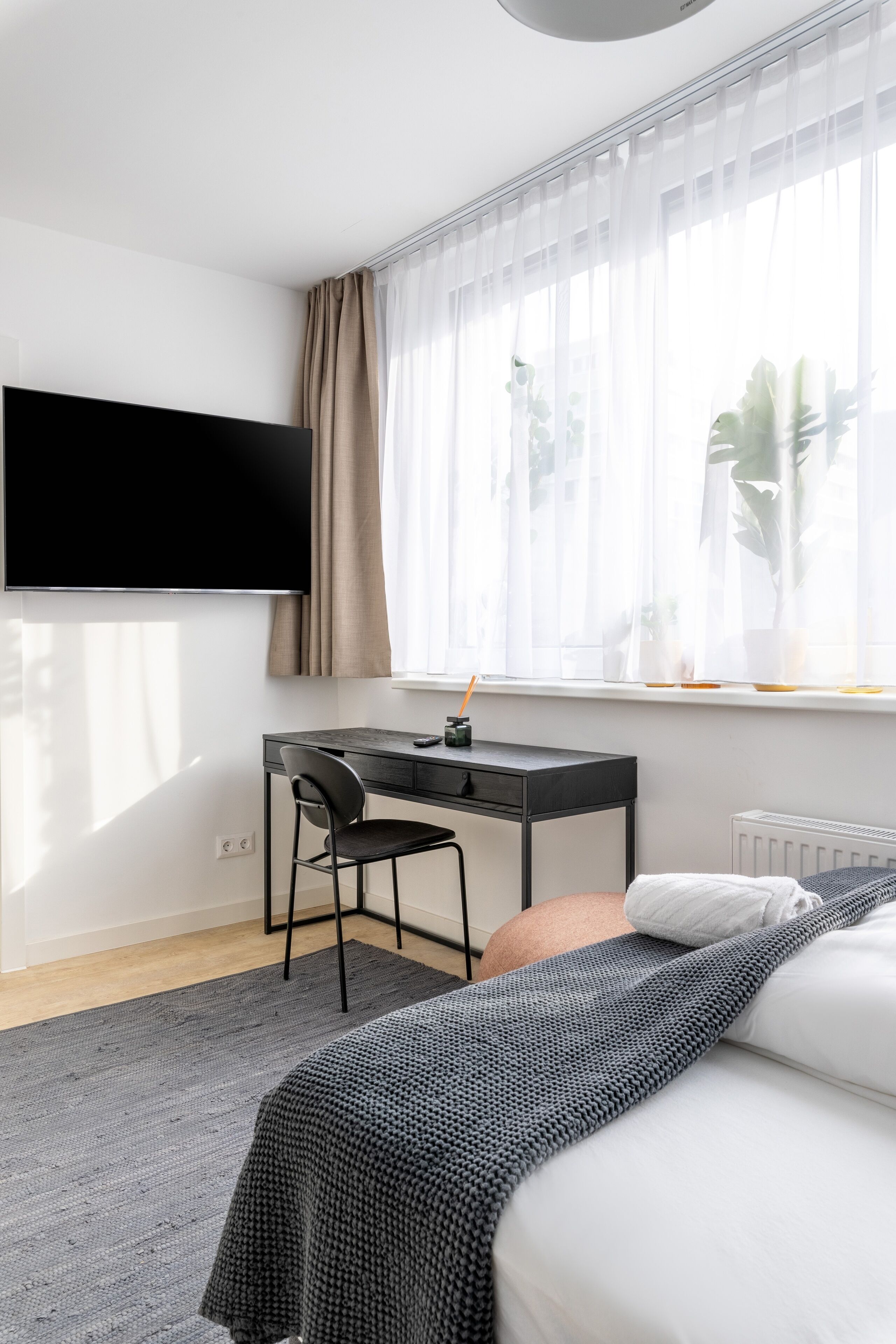 Ein modernes Hotelzimmer mit einem großen Flachbildfernseher an der Wand, einem Schreibtisch mit Stuhl, einem Bett mit grauer Decke und einem Fenster mit transparenten Vorhängen.