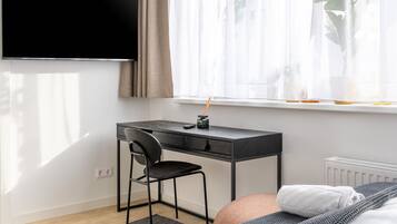 Ein modernes Hotelzimmer mit einem großen Flachbildfernseher an der Wand, einem Schreibtisch mit Stuhl, einem Bett mit grauer Decke und einem Fenster mit transparenten Vorhängen.
