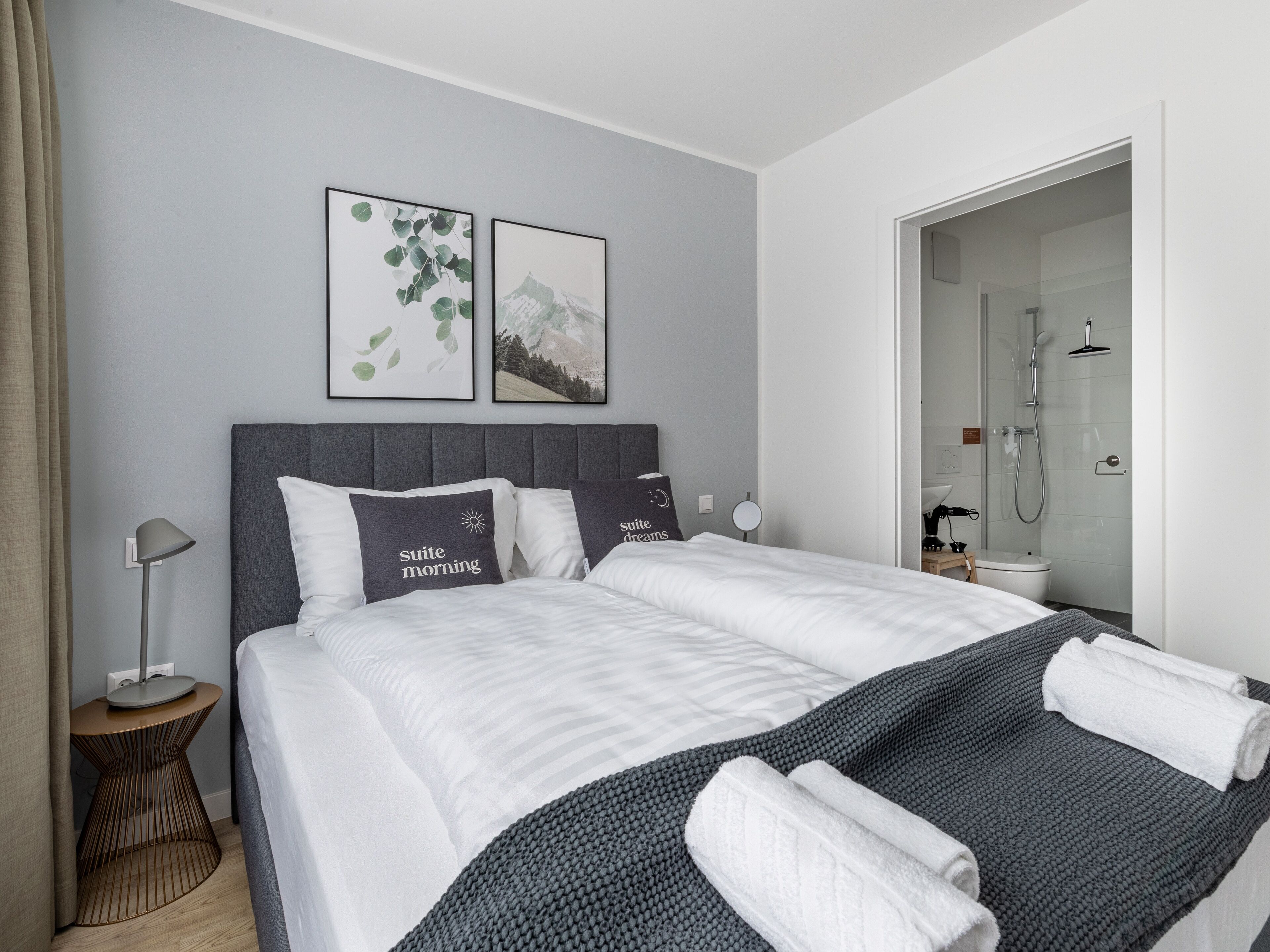 Quarto de hotel com uma cama grande, duas almofadas com texto, uma mesa de cabeceira, um abajur e área de chuveiro.