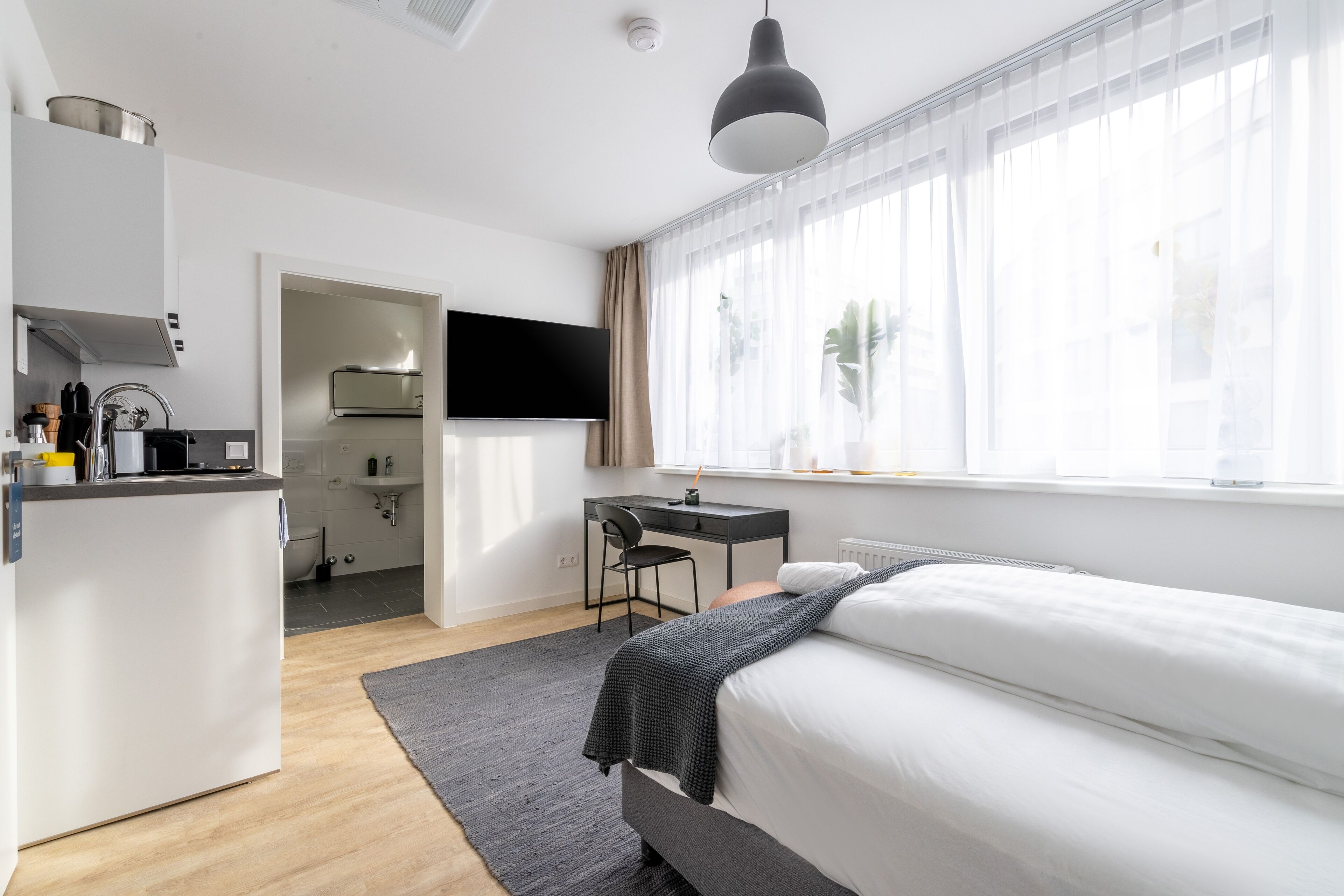 A modern hotel room with a bed, a desk, and a kitchenette.