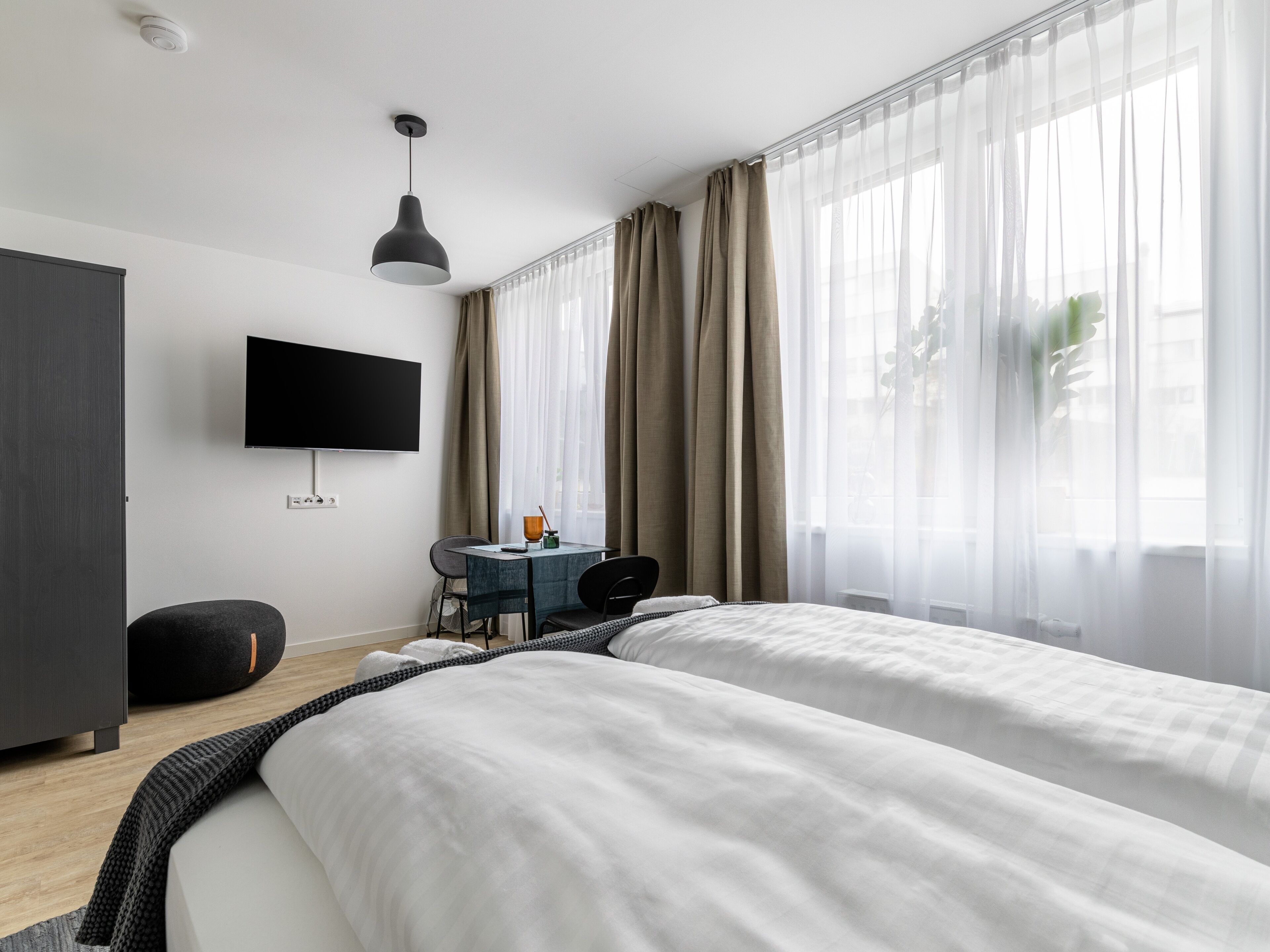 A modern hotel room with two beds, a desk, a chair, a TV, and sheer curtains.