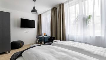 A modern hotel room with two beds, a desk, a chair, a TV, and sheer curtains.