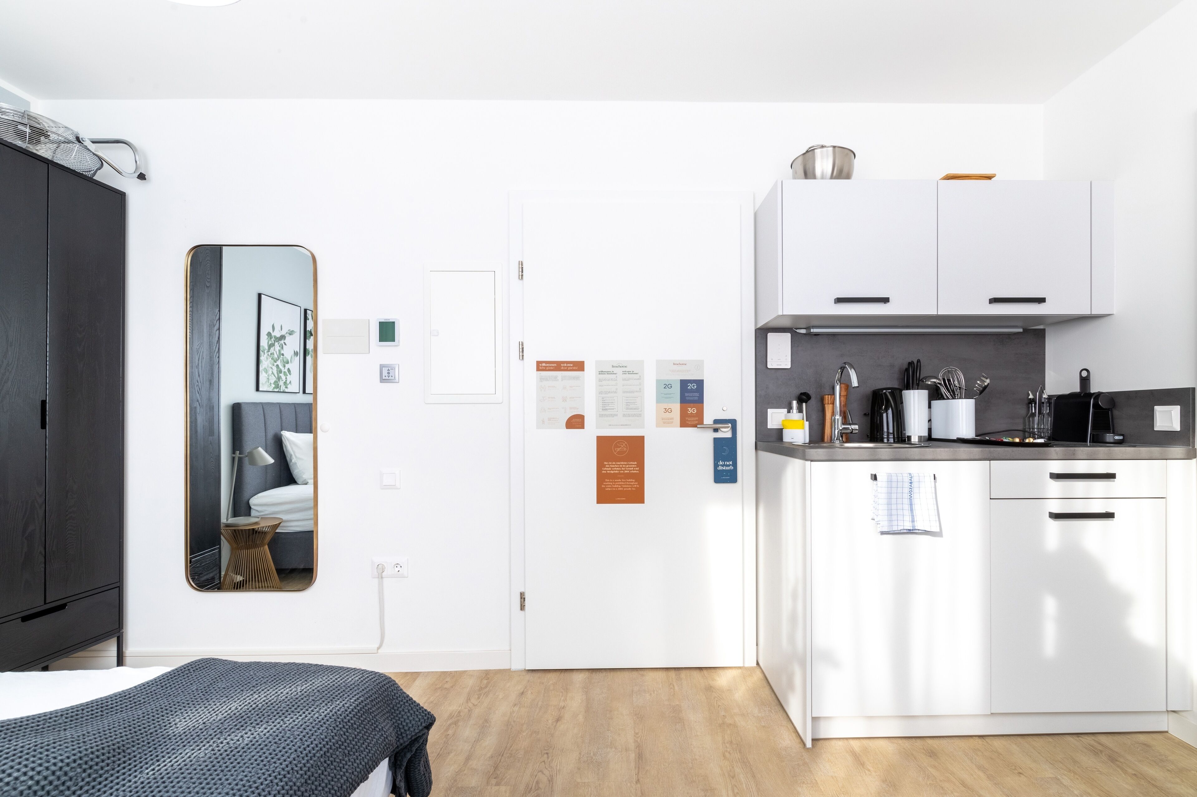 A modern hotel room with a bed, a mirror, a small side table, a kitchenette with white cabinets and a countertop, and a door with informational cards.