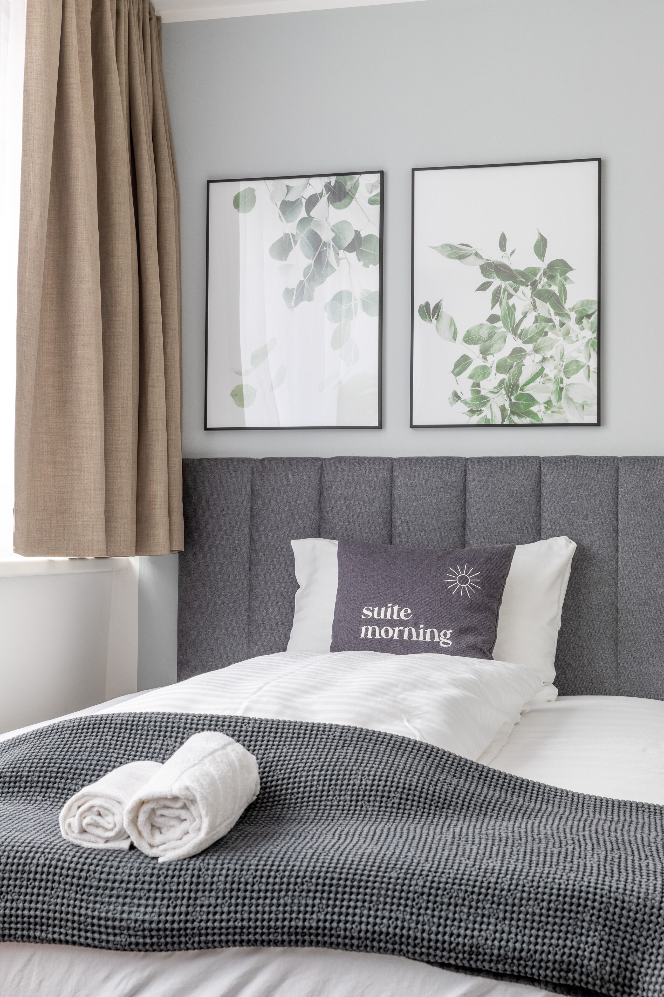 Ein ordentlich bezogenes Bett mit grauem Kopfteil, einer grauen und weißen Decke und zwei aufgerollten Handtüchern. An der Wand über dem Bett hängen zwei gerahmte Blätterbilder.