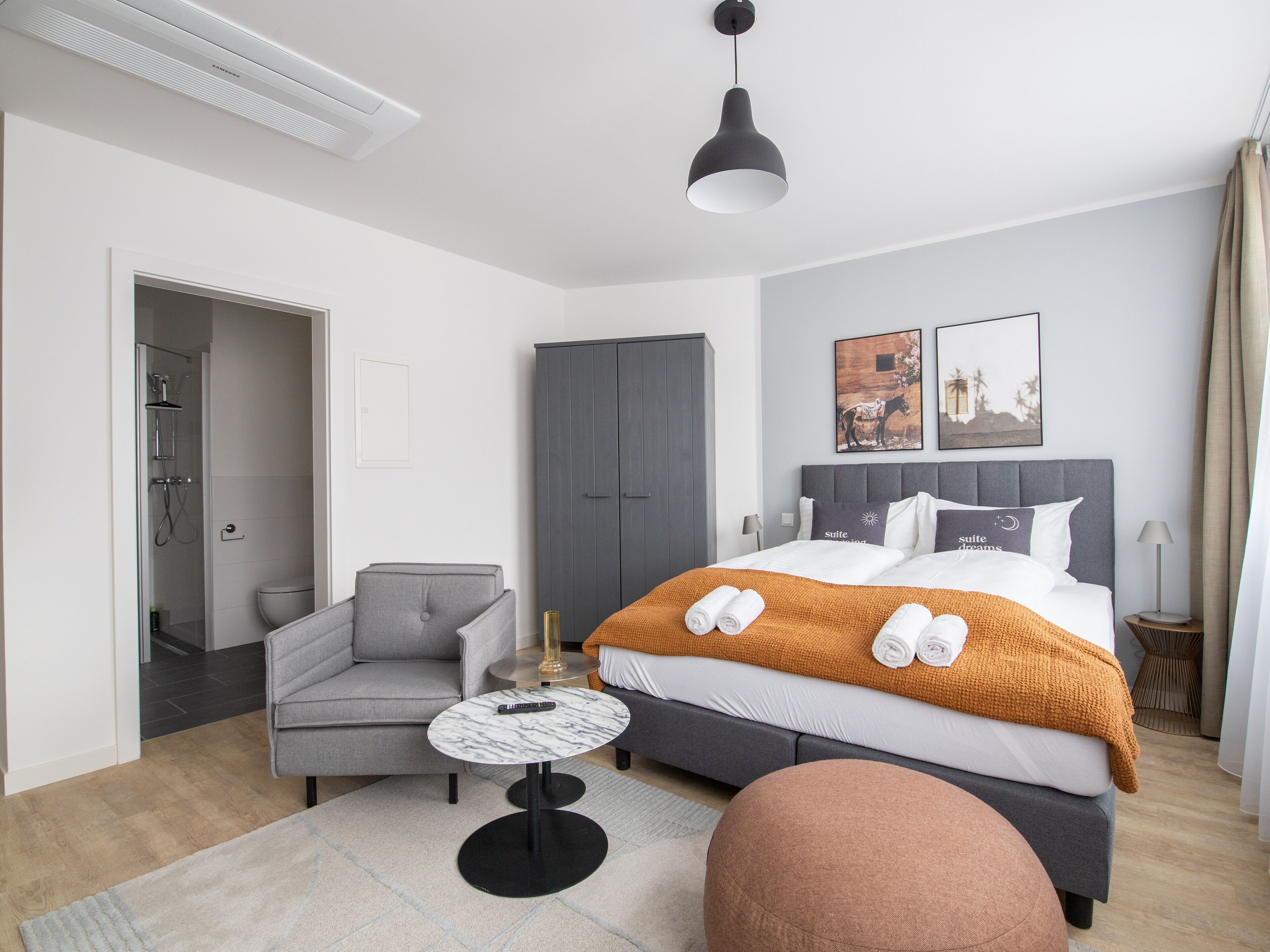 Une chambre d’hôtel moderne comprenant un lit, un fauteuil gris, une petite table ronde, un espace douche et une armoire.