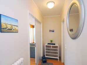 A hallway with a wooden floor, a white radiator, a mirror, a small plant on a white dresser, and a painting of a beach scene.