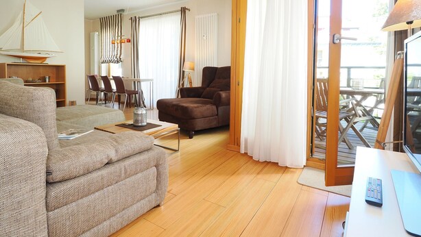 A living room with a sofa, armchair, dining area, and a view through a sliding door to an outdoor deck. - Apartment with 1 bedroom and balcony (Seebad Heringsdorf)