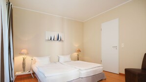 A double bed with white bedding, two bedside lamps, a painting of sailboats, a wooden floor, and a door. - Apartment with 1 bedroom and balcony (Seebad Heringsdorf)