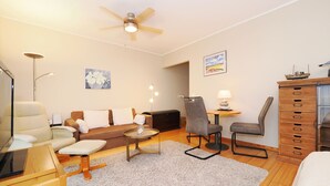 A well-lit, spacious room with a ceiling fan, a sofa, a chair, a small table with a lamp, a wooden cabinet, and a painting on the wall. - 1-room apartment with terrace (Seebad Heringsdorf)